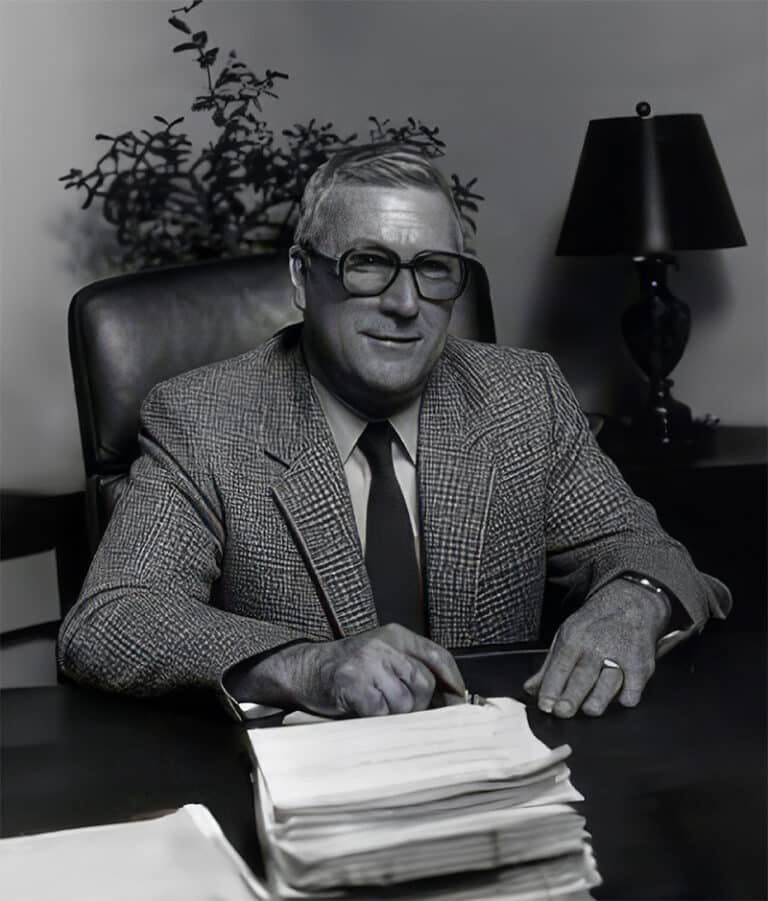 A man wearing glasses seated at a desk with documents.