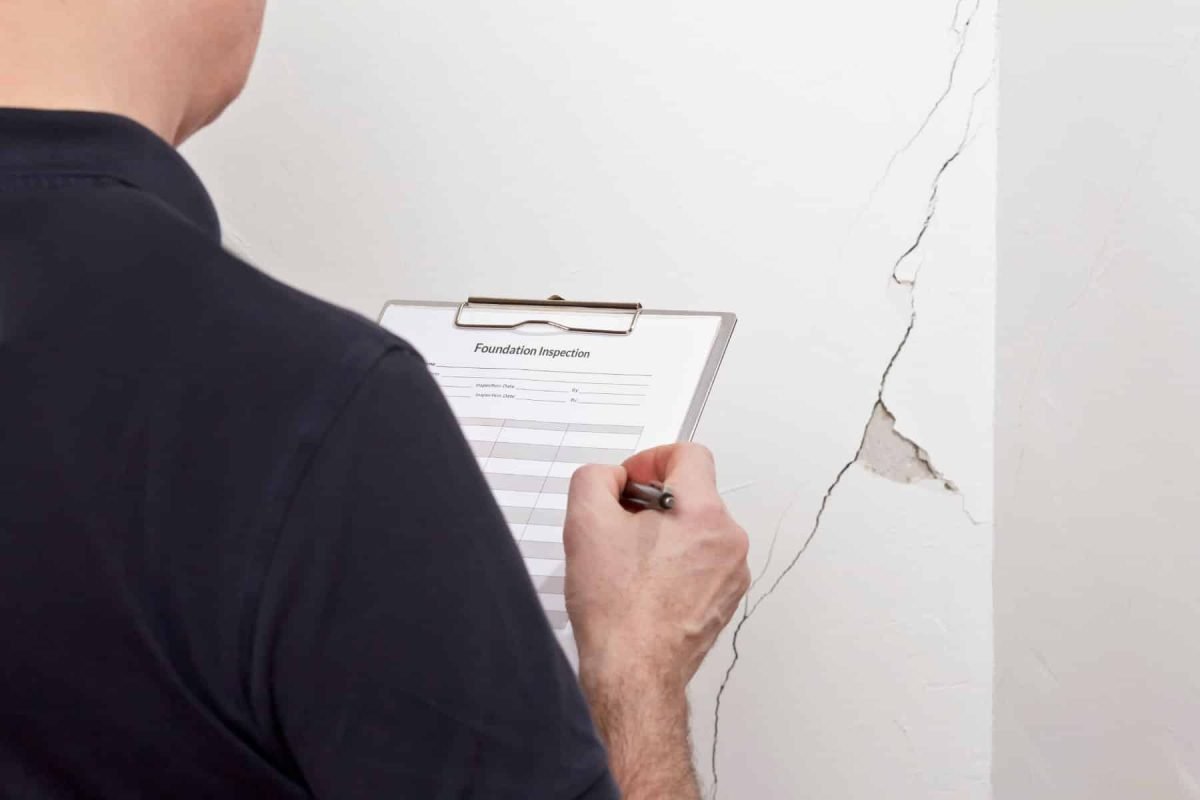 Man with inspection checklist in front of a white wall with a long crack or rip and a piece of plaster missing.